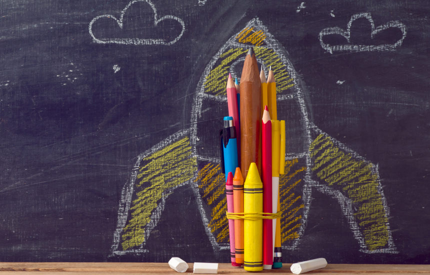 Photo of a rocket drawn on a chalkboard and pencils/crayons with a rubber band around them in front of it.