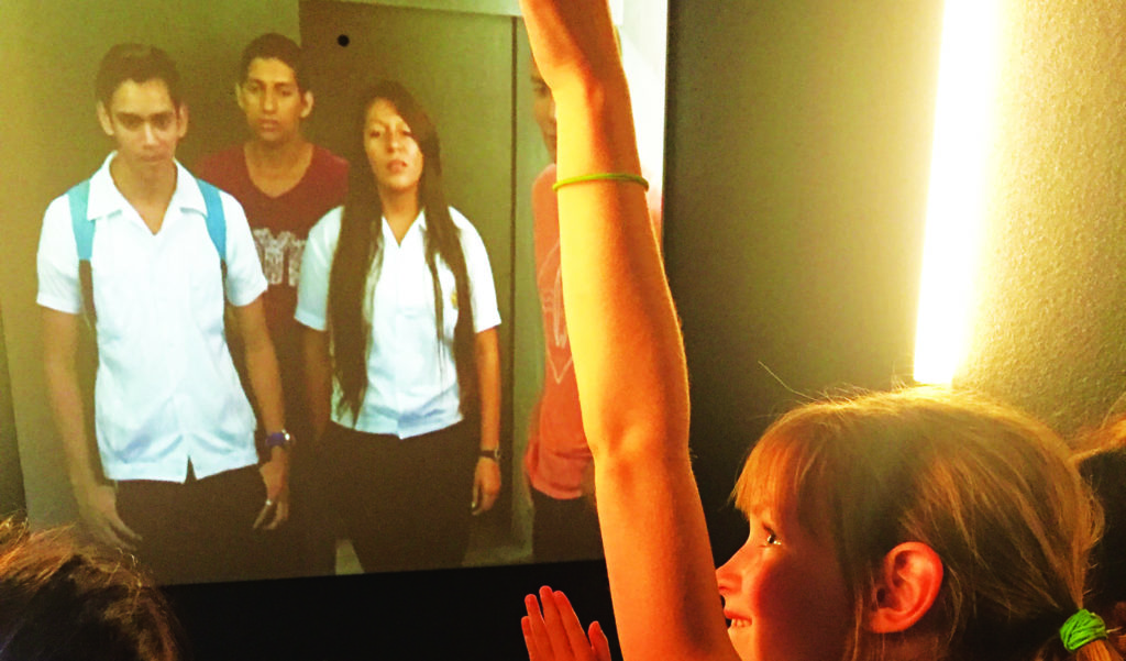 A young girl raises her hand in front of a video screen