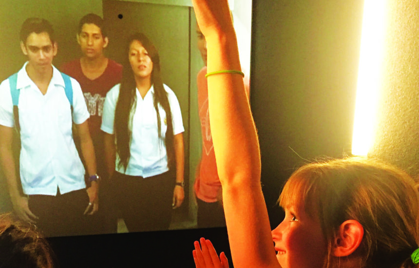 A young girl raises her hand in front of a video screen