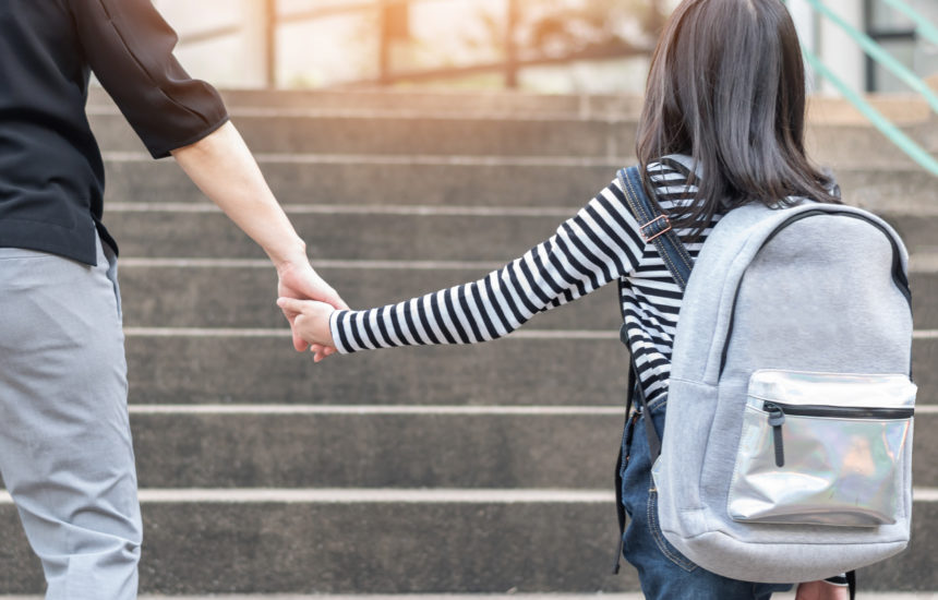 An adult holds the hand of a young girl walking up steps wearing a backpack