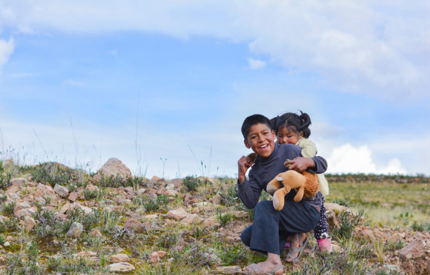 A young boy smiles and carries a young girl holding a stuffed animal on his back outside