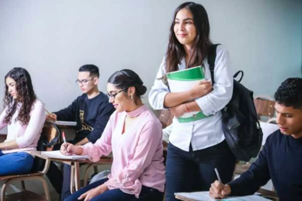 A woman wearing a backpack and carrying a textbook walks into a classroom of people taking notes