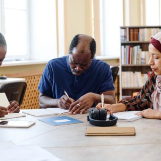 Photo of two women and one man writing on papers on a desk in a library