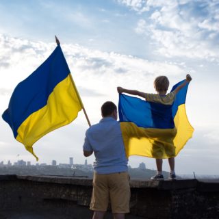 Photo of a dad and child holding Ukrainian flags, looking out at a city in the distance