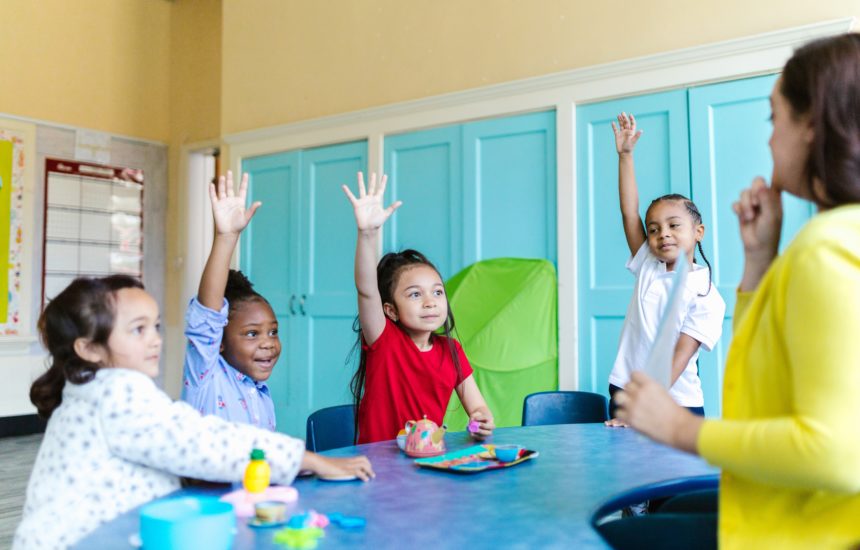 Photo of a teacher surrounded by young kids excitedly raising their hands