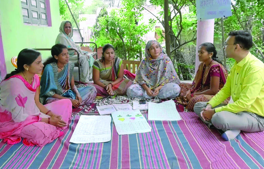 Six people talking while sitting in a semi circle. One smiling woman in the background looking at group