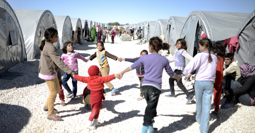 Children in a refugee camp who are playing a game where they walk in a circle, holding hands. In the background are rows of temporary housing tents.