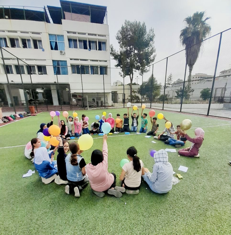 Photo of kids sitting in a schoolyard in a circle, holding balloons