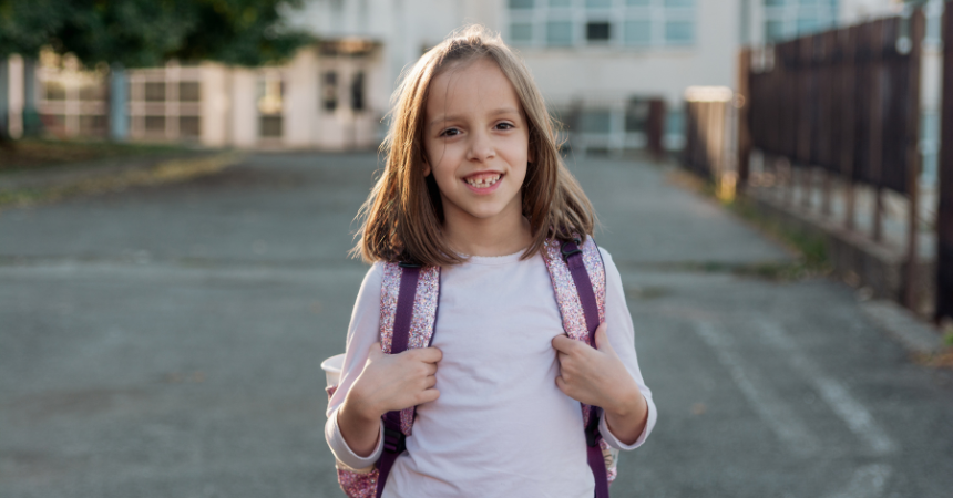 A young girl with a backpack in front of a school in Serbia, smiling