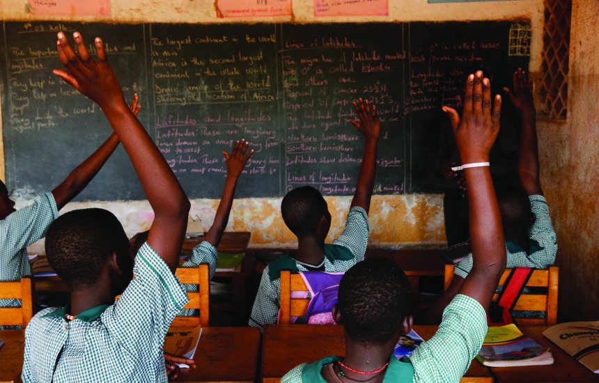 Children in Mulago school for the deaf in Uganda, facing away from the camera towards a blackboard with writing on it. They are raising their hands to be called on by a teacher. The students have dark-skin and short black hair; they are wearing matching blue uniforms.