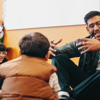 A teacher sitting on the floor with elementary students, happily explaining something to them.