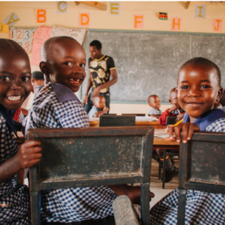 Three happy girls sitting in desks in a classroom with uniforms on. Their teacher in the background is helping another student.