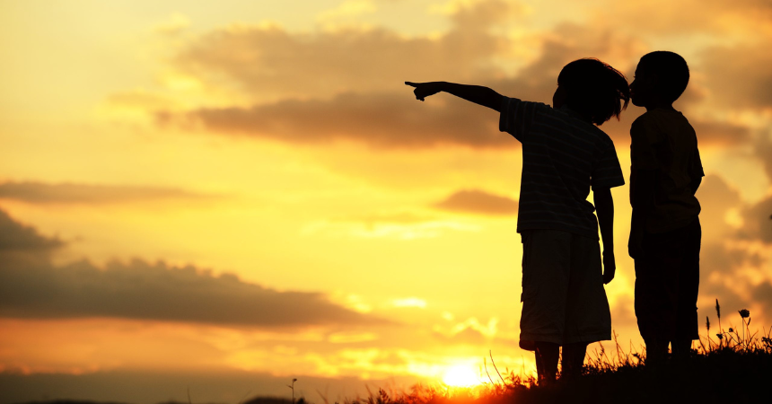 Two children standing together at sunset, with one of them pointing to something in the distance to show something to the other.