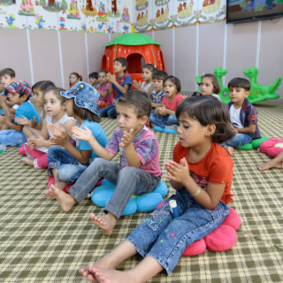 Happy young children sitting on the floor in a classroom, singing and making hand movements.