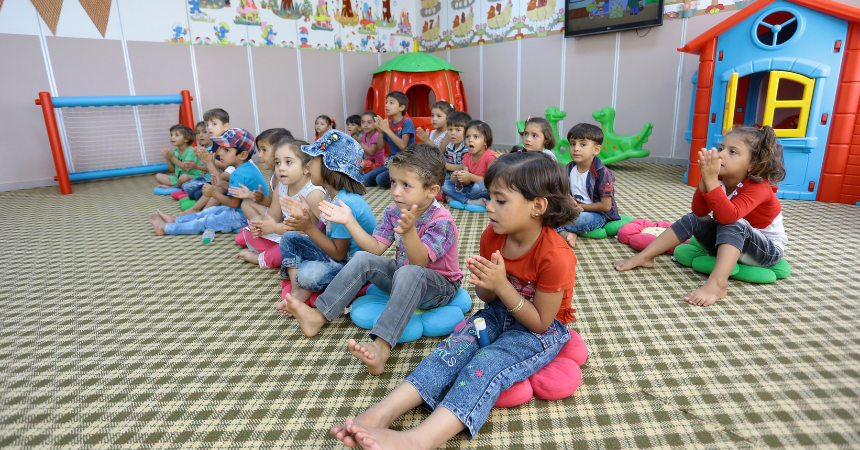 Happy young children sitting on the floor in a classroom, singing and making hand movements.