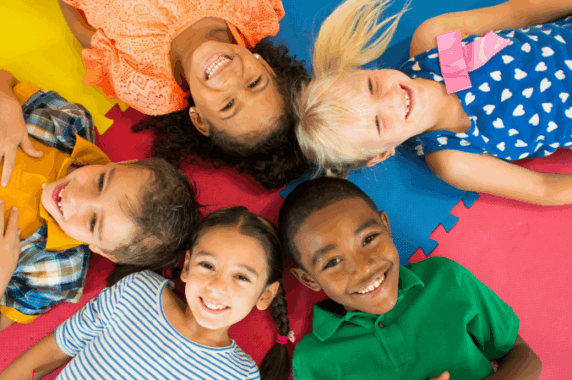 Five smiling children lying together in a circle on a colorful mat, smiling.
